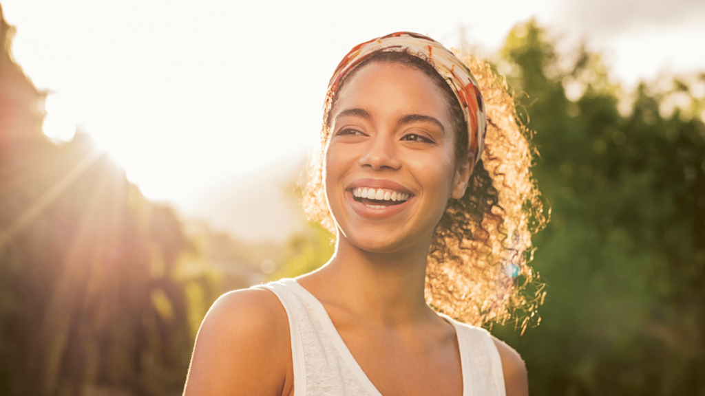 Uma mulher negra que sorri e tem um lenço amarrado no cabelo está no foco da imagem. Ao fundo, há um ambiente de natureza e o reflexo da luz do sol.