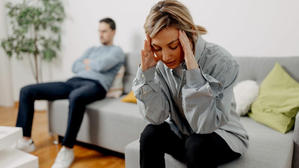 Imagem de um casal, sentandos em um sofá. Em destaque, a imagem de uma mulher com as mãos sobre a cabeça, trazendo o conceito de exaustão, de cansaço, de problemas no relacionamento.
