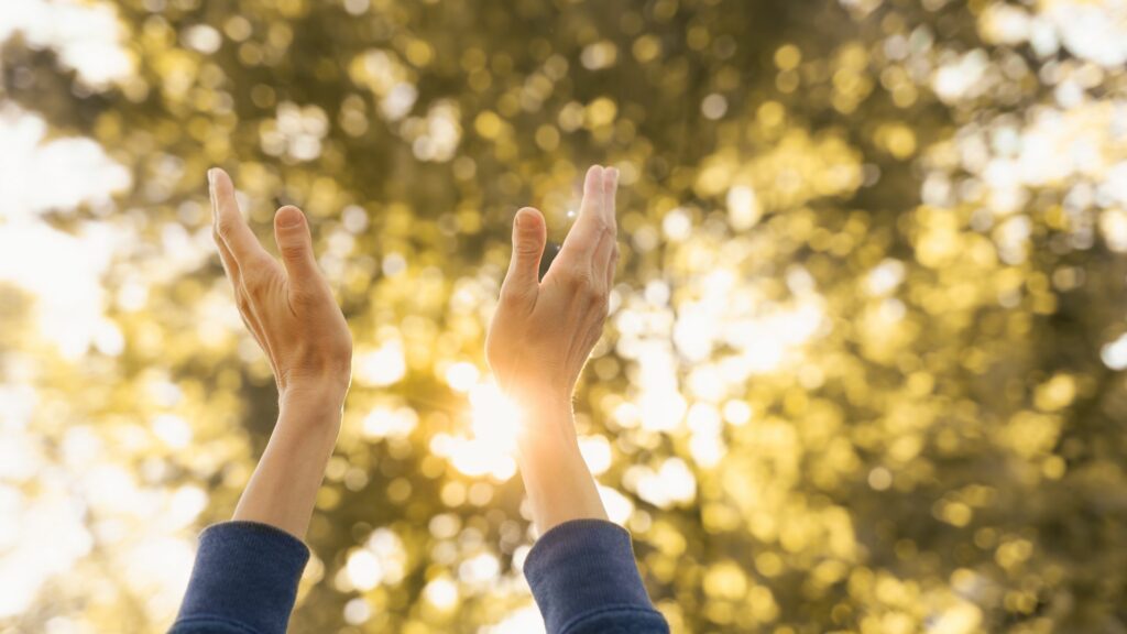 Imagem das mãos de uma mulher erguidas para o céu durante o pôr do sol. A imagem traz o conceito de energia positiva, oração e gratidão.
