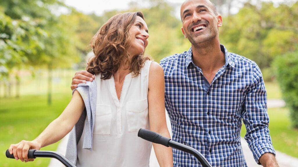 Imagem de um casal maduro e feliz, caminhando em um parque ao lado de suas bicicletas.
