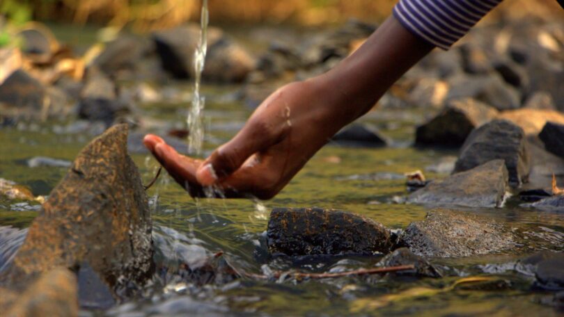 Imagem de uma fonte de água natural e limpa e a mão de uma pessoa tocando nesta água.