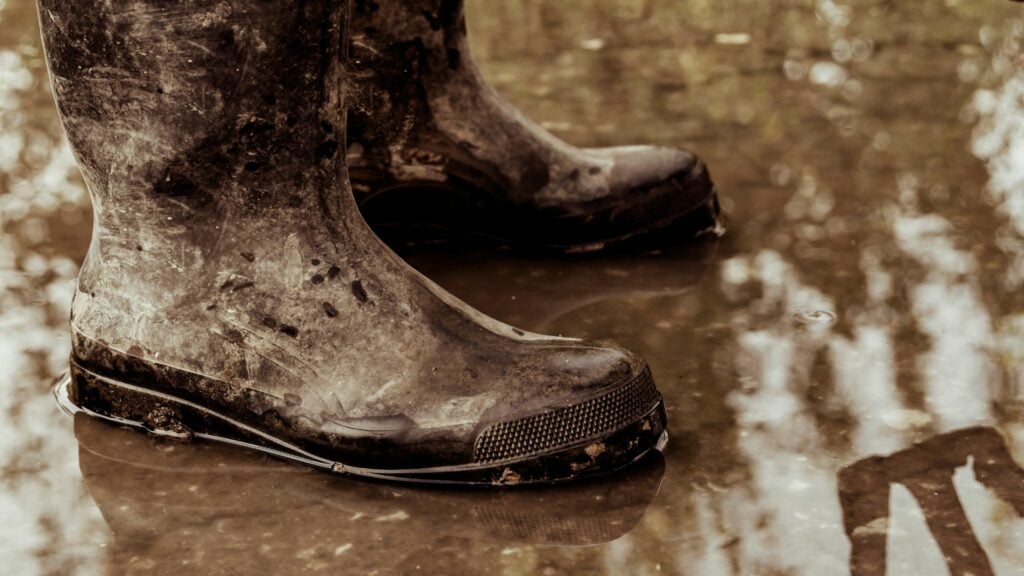 Imagem de um pé usando botas sujas na lama e na água suja.
