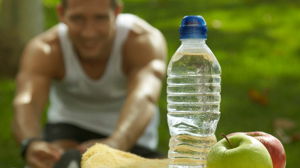 Imagem de um homem fazendo atividade física em uma praça e em destaque uma garrafa de água e duas maçãs, trazendo o conceito de nutrição e hidratação após o esporte.
