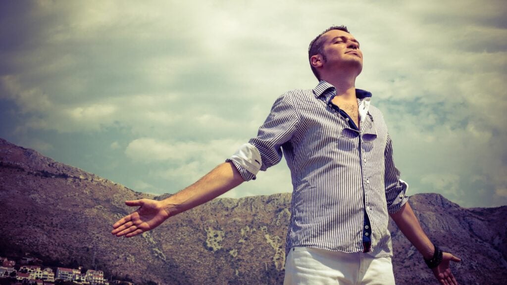 Imagem de um homem, usando uma camisa listrada nas cores azul e branco, uma calça branca. Ele está de braços aberto desfrutando de liberdade e ar fresco para seguir a vida.