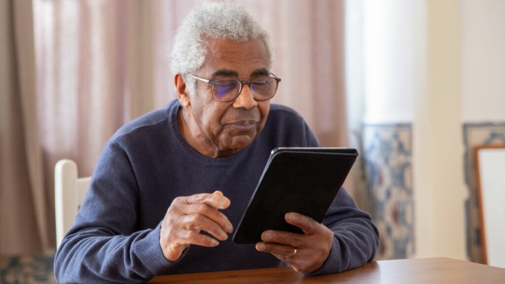 Um homem idoso de suéter azul usando um tablet digital, trazendo o conceito de desafios com a tecnologia e a terceira idade.
