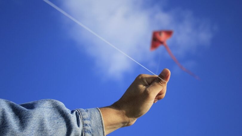 Imagem de um céu azul e em destaque uma pipa no céu e o braço de uma pessoa segurando a linha, trazendo o conceito de libertade e autonomia.